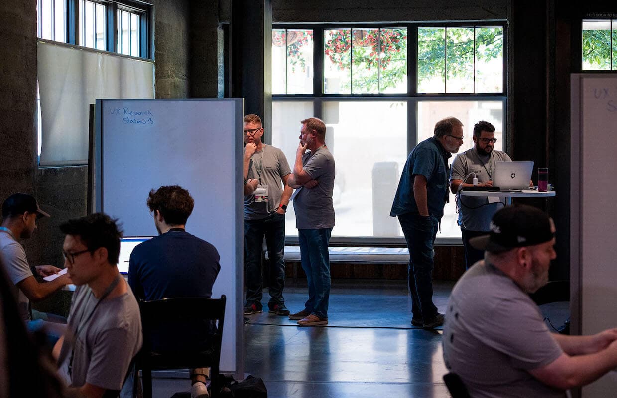 Different people gathering in small groups to discuss identity architecture solutions using a laptop or whiteboard.