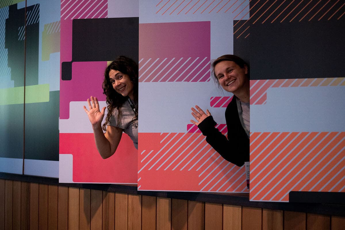 Two people peaking through decorative wall panels to smile and wave.
