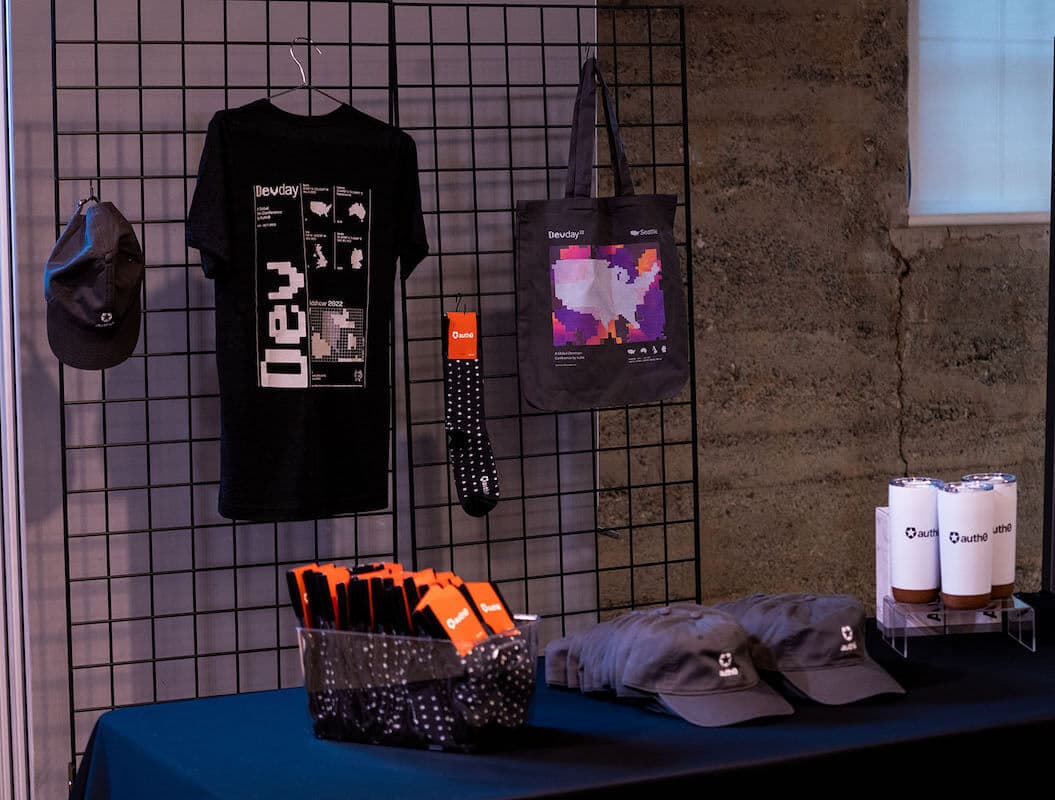 Items given at DevDay22 for swag displayed over a table and a wall fence, including a cap, tee, a pair of socks, a totem bag, and thermal cups.